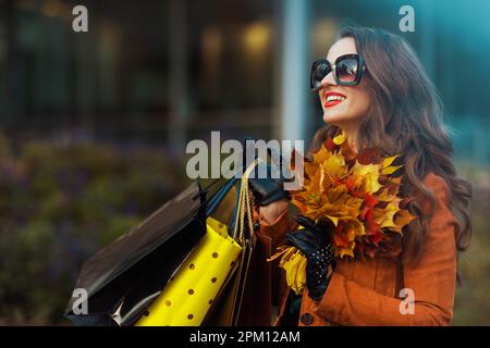 Hallo Herbst. Glückliche, moderne 40-jährige Frau in orangefarbenem Trenchcoat mit Einkaufstaschen und herbstgelben Blättern in der Nähe des Geschäfts in der Stadt. Stockfoto