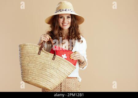 Strandurlaub. Lächelnde moderne 40 Jahre alte Hausfrau in weißer Bluse und Shorts vor beigefarbenem Hintergrund mit Verbandskasten, Strohtasche und Sommerhut Stockfoto