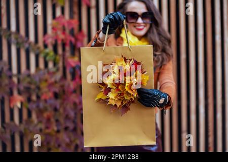 Hallo Herbst. Nahaufnahme einer glücklichen, eleganten Frau mittleren Alters in orangefarbenem Trenchcoat mit Einkaufstasche und herbstgelben Blättern in der Stadt. Stockfoto