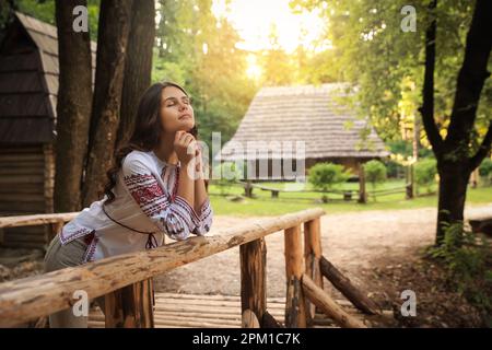 Schöne Frau mit gesticktem Hemd in der Nähe des Holzgeländers auf dem Land, Platz für Text. Ukrainische Nationalkleidung Stockfoto