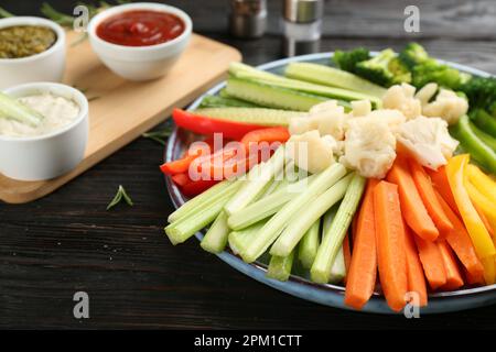 Teller mit Selleriestangen, anderem Gemüse und verschiedenen Dip-Saucen auf dunklem Holztisch, Nahaufnahme Stockfoto