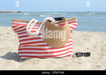 Stilvolle gestreifte Tasche mit Visierkappe und Sonnenbrille am Sandstrand in der Nähe des Meeres Stockfoto