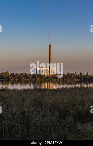 Eine alte Müllverbrennungsanlage am Seeufer mit rauchenden Schornsteinen vor einem bunten Himmel. Die Energieerzeugung verursacht Umweltverschmutzung. Stockfoto