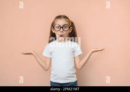 Süßes, lustiges, kleines, weißes Mädchen mit runden Gläsern kann nicht auswählen, was im leeren Raum ist, wenn es auf einen rosa Hintergrund in die Kamera schaut Stockfoto