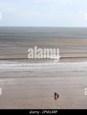 An einem Frühlingsmorgen brechen die Wellen sanft an die Küste, während ein Paar mit seinem Jungen in einem Liegestuhl entlang des ruhigen Hunmanby Sands spaziert Stockfoto