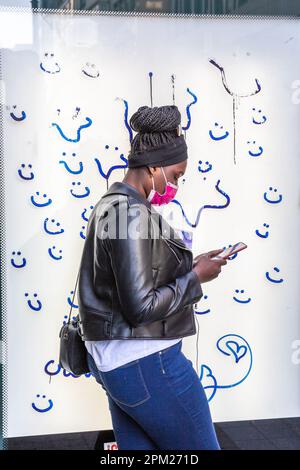 Junge schwarze Frau, die eine Sanitärmaske trägt und ihr Handy konsultiert, vor dem markierten Schild einer Busunterkunft. Brüssel. Stockfoto