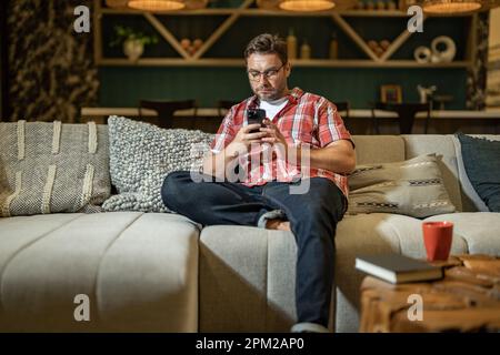 Freiberuflicher Geschäftsmann, der sein Mobiltelefon verwendet, um zu Hause soziale Medien oder Nachrichten-Chats zu überprüfen. Online arbeiten von zu Hause aus. Ein Mann ruht sich nach einem langen Arbeitstag aus Stockfoto