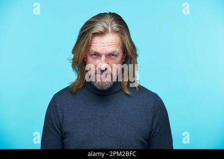 Porträt eines reifen bärtigen Mannes, der in die Kamera schaut, mit wütendem Ausdruck isoliert auf blauem Hintergrund Stockfoto