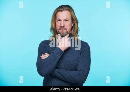 Porträt eines erwachsenen Mannes mit langen Haaren, der in die Kamera schaut, mit pensivem Ausdruck auf blauem Hintergrund Stockfoto