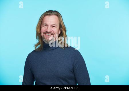 Porträt eines gut aussehenden, erwachsenen Mannes mit langen Haaren, die vor der Kamera lächeln und auf blauem Hintergrund stehen Stockfoto