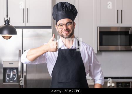 Glücklicher Bäcker mit Daumen nach oben. Porträt eines Koch-Mannes in einer Kochmütze in der Küche. Mann mit Schürze und Kochuniform und Köchmütze. charakterküche Stockfoto