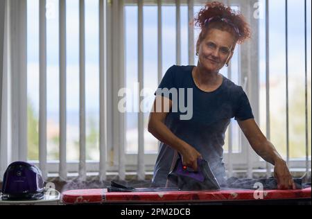 Lockiges Haar, rothaarige Hausfrau, die Kleider am Fenster bügelt Stockfoto