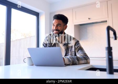 Mann Zu Hause, Der An Einem Laptop An Der Theke In Der Küche Arbeitet Stockfoto