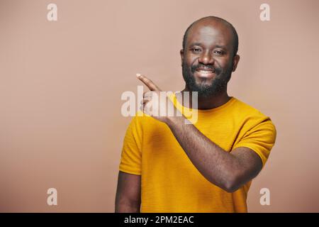 Porträt eines afroamerikanischen bärtigen Mannes, der auf leeren Raum zeigt und vor der Kamera lächelt, auf braunem Hintergrund Stockfoto