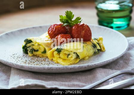 Hausgemachtes Omelett mit Spinat und gerösteten Kirschtomaten auf Keramikplatte auf rustikalem Holztisch, gesundes französisches Frühstücks-Omelette mit Grünpflanzen Stockfoto