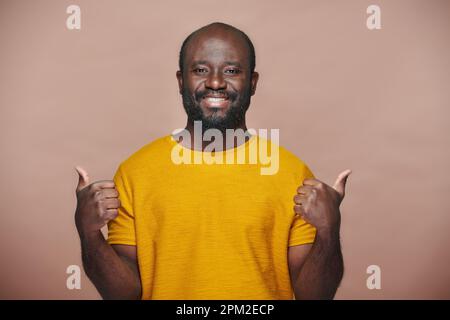 Porträt eines afroamerikanischen Mannes in gelbem T-Shirt, der Daumen hoch zeigt und vor der Kamera lächelt Stockfoto