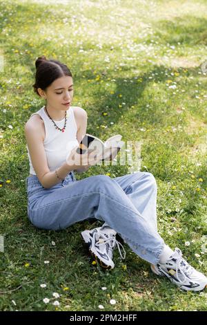 Blick von oben auf die junge brünette Frau und Jeans, die Buch lesen, während sie auf der Wiese mit Blumen im Park sitzen, Stockbild Stockfoto