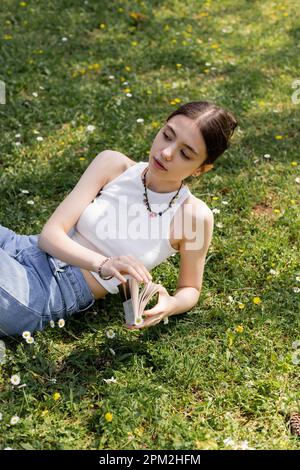 Blick von oben auf junge brünette Frau und Jeans mit Buch, während sie auf dem Rasen liegen und Gänseblümchen im Park, Stockbild Stockfoto