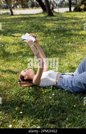 Junge brünette Frau mit Oberteil und Jeans, die Buch lesen, während sie auf der Wiese liegt, mit Blumen im Park, Stockbild Stockfoto