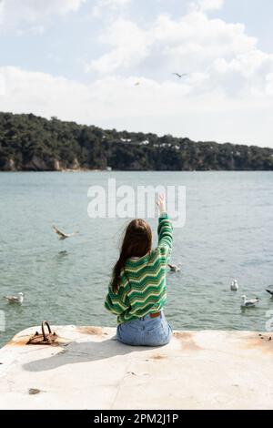 Rückansicht einer jungen Frau, die auf dem Pier neben verschwommenen Möwen auf dem Wasser in der Türkei sitzt, Stockbild Stockfoto