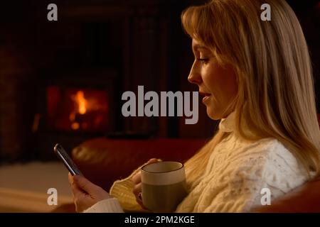 Frau Zu Hause In Der Lounge Mit Gemütlichem Feuer Und Mobiltelefon Stockfoto