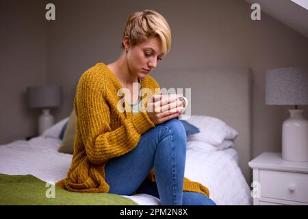 Unglückliche Junge Frau Mit Heißem Getränk, Die Zu Hause Auf Dem Bett Sitzt Stockfoto