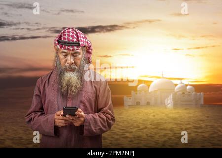 Ein muslimischer Mann mit Bart, der Keffiyeh mit Agial trägt und ein Mobiltelefon mit dem Hintergrund der Sonnenuntergangsszene benutzt Stockfoto