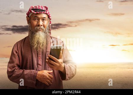 Ein muslimischer Mann mit Bart, der Keffiyeh mit Agial trägt und ein Mobiltelefon mit dem Hintergrund der Sonnenuntergangsszene benutzt Stockfoto