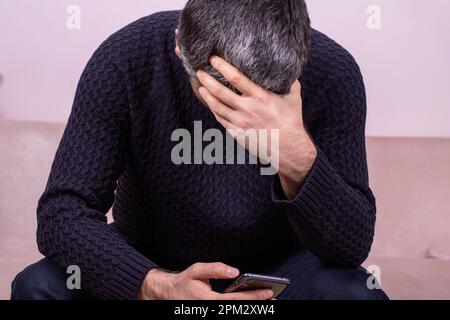 Ein Mann mittleren Alters, der am Telefon traurige Nachrichten erhält. Besorgter Mann liest Nachrichten online, sieht auf den Telefonbildschirm, weiß sitzt zu Hause auf dem Sofa, ernst Stockfoto