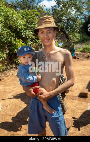 Ein junger Vater aus einer ethnischen Minderheit Jerais mit einem großen Tattoo trägt seinen Sohn in den zentralen Highlands Vietnams. Stockfoto