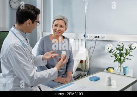 Junger Arzt im weißen Kittel, der Patienten Medikamente zur Behandlung verschreibt, während sie auf der Station auf dem Bett sitzen Stockfoto
