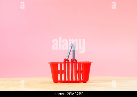Leerer roter Warenkorb auf rosa Hintergrund, Supermarkt Hintergrund für Lebensmitteleinkäufe auf Holztisch. Helles Geschäft mit Produkten und Speisen im Flur Stockfoto