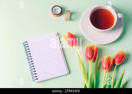 Spiralnotizblock, eine Tasse Tee und rote Tulpenblüten auf hellgrünem Hintergrund. Draufsicht, flach liegend, Kopierbereich. Stockfoto