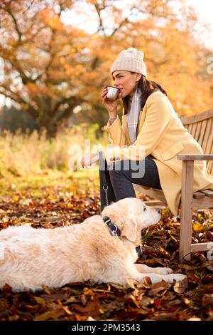 Frau Mit Hut Und Schal Mit Heißem Getränk Sitzt Auf Einer Bank Mit Hund Auf Einem Spaziergang In Der Herbstlandschaft Stockfoto