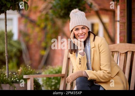 Porträt Einer Frau Mit Hut Und Schal, Die Im Herbst Auf Der Bank Vor Dem Haus Sitzt Stockfoto