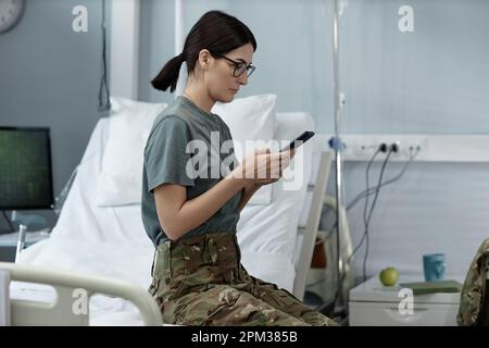 Junge Soldatin tippt Nachricht auf Smartphone, während sie auf der Station auf dem Bett sitzt Stockfoto