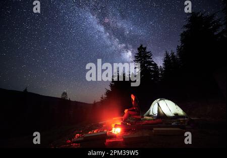 Nächtlicher Campingausflug in den Bergen, inmitten funkelnder Sterne. Eine touristische Frau ruht am Lagerfeuer und im Zelt, eingebettet in der Nähe des Waldes, unter dem prächtigen Baldachin der Milchstraße, unter dem faszinierenden Nachthimmel. Stockfoto