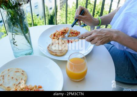 Eine unbekannte Frau, die Arepas isst, Rühreier mit Orangensaft, auf dem Balkon einer Wohnung, wo sie ein ruhiges Frühstück bekommt. Lifestyle c Stockfoto