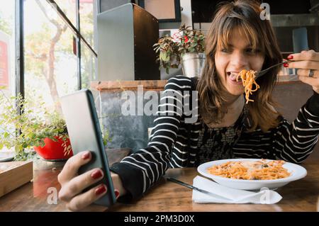 Junge transsexuelle argentinische latinin mit langen Haaren und weißer Haut, mit roten Nägeln, sitzt in einem Restaurant und isst Spaghetti, während sie sich einen holt Stockfoto