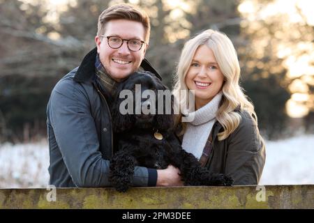 Portrait Eines Liebevollen Paares Mit Haustier Spaniel Dog Leaning On Fence On Snowy Walk In Winter Countryside Stockfoto