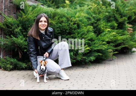 Frau in einer legeren Jacke, die am Herbsttag in der Nähe ihres Lieblingshundes sitzt, der in einem Anzug für Hunde auf der Straße trainiert ist Stockfoto