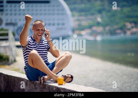Ein junger Mann spricht auf dem Smartphone, während er sich an einem schönen sonnigen Tag während eines Urlaubs am Meer am Kai ausruht. Urlaub, am Meer, Stockfoto
