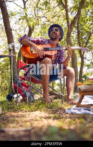 Ein lächelnder Mann, der in der Natur zeltet und Gitarre spielt Stockfoto