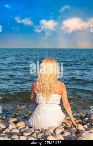 Frau in weißem Kleid, von hinten gesehen, sitzt an einem Kieselstrand und blickt aufs Meer. Stockfoto