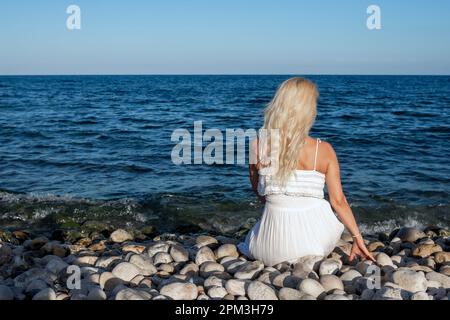 Blonde Frau in weißem Kleid, von hinten gesehen, sitzt an einem Kieselstrand und blickt aufs Meer. Stockfoto