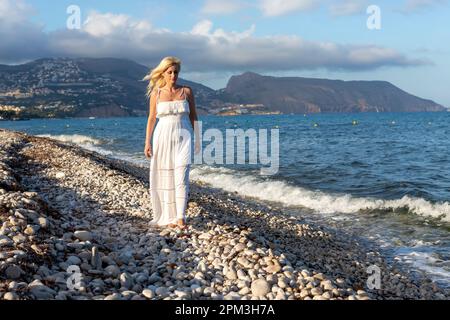 Eine blonde Frau in weißem Kleid, die am Kieselstrand spaziert, bei Sonnenuntergang Stockfoto