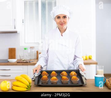 Lächelnde Köchin in weißer Uniform, die süße Cupcakes backt Stockfoto