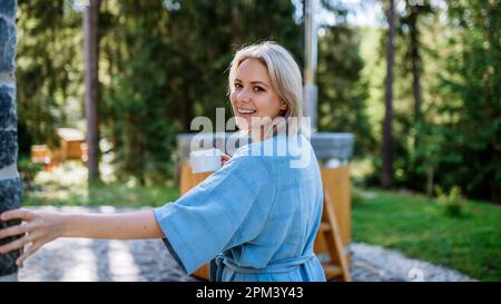 Glückliche junge Frau, die im Garten neben dem Whirlpool steht und im Sommerurlaub in den Bergen eine Tasse Kaffee am Morgen genießt. Stockfoto