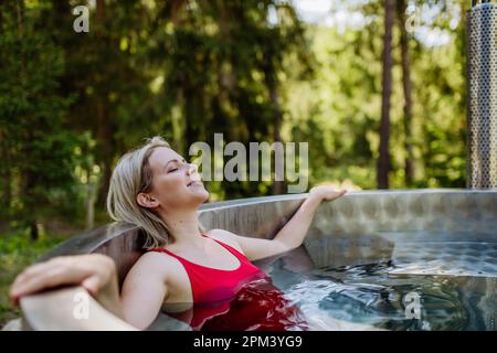 Junge Frau, die in der Holzbadewanne mit Kamin Holz verbrennt und Wasser im Hinterhof in den Bergen erwärmt. Stockfoto