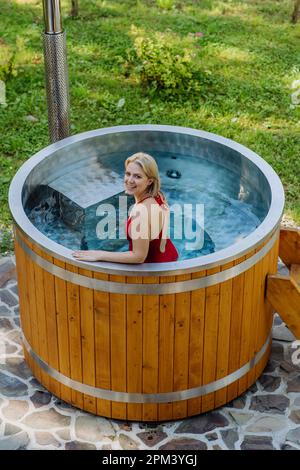 Junge Frau, die in der Holzbadewanne mit Kamin Holz verbrennt und Wasser im Hinterhof in den Bergen erwärmt. Stockfoto